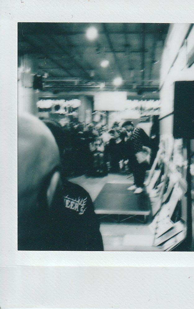 The image depicts a crowded indoor event with people gathered around a small stage in a dimly lit setting.