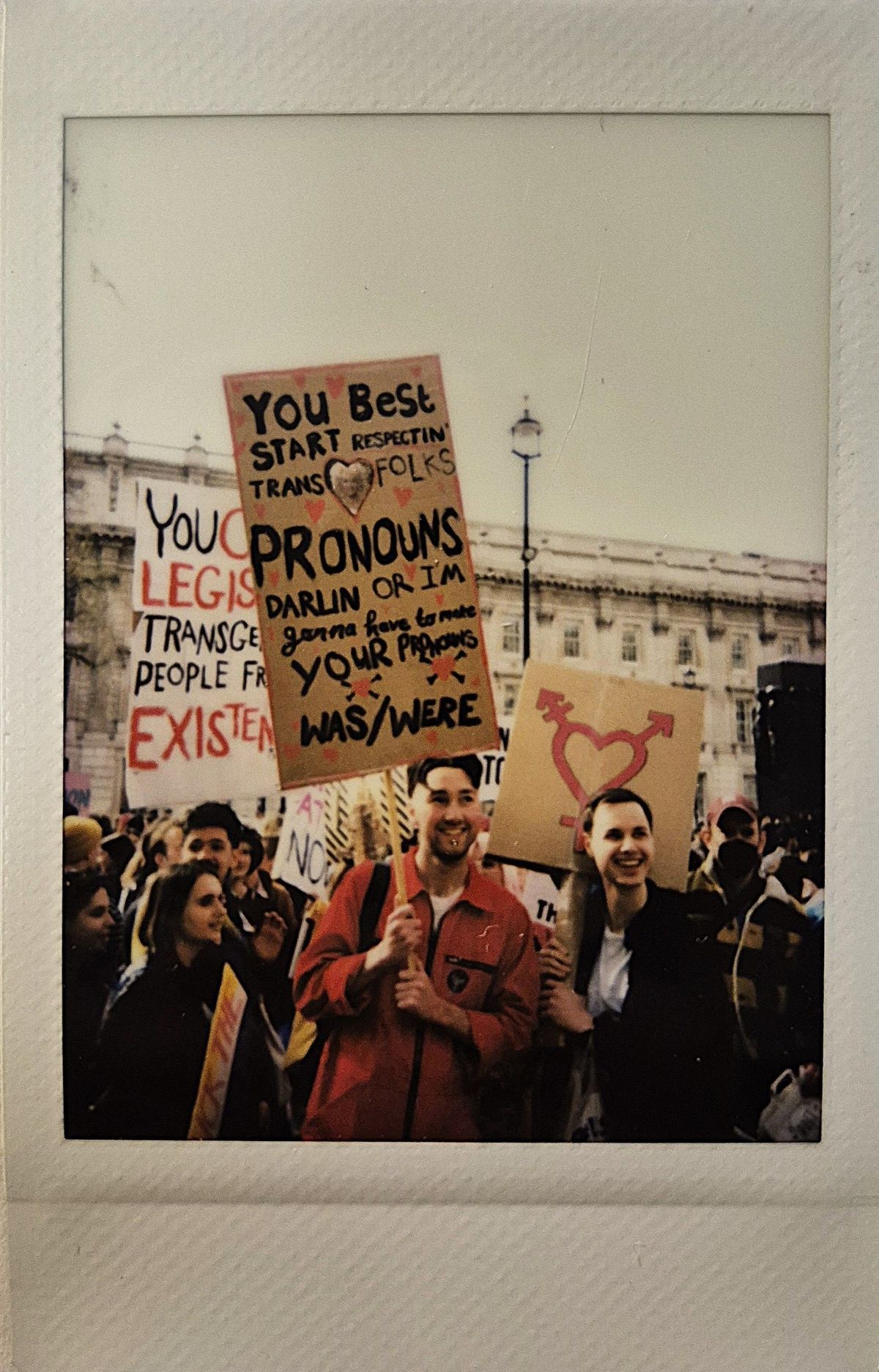 A group of people holding protest signs, one says "YOU BEST START RESPECTING TRANS FOLKS PRONOUNS DARLIN OR I'M GONNA HAVE TO MAKE YOUR PRONOUNS WAS/WERE".