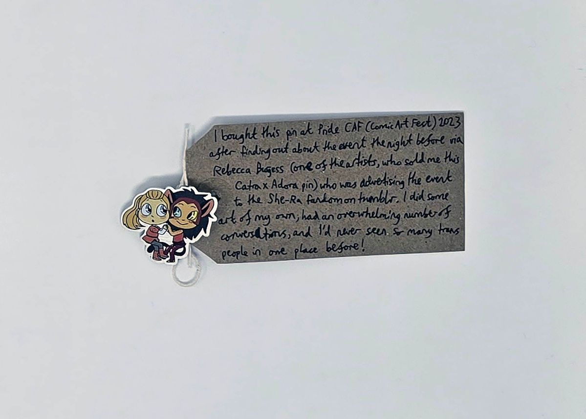 An enamel pin showing two people embracing and holding hands. One is blonde and wearing a red outfit and the other has brown hair and animal ears. They are characters from the cartoon She-Ra. The pin is attached to a brown tag with a handwritten message on it.