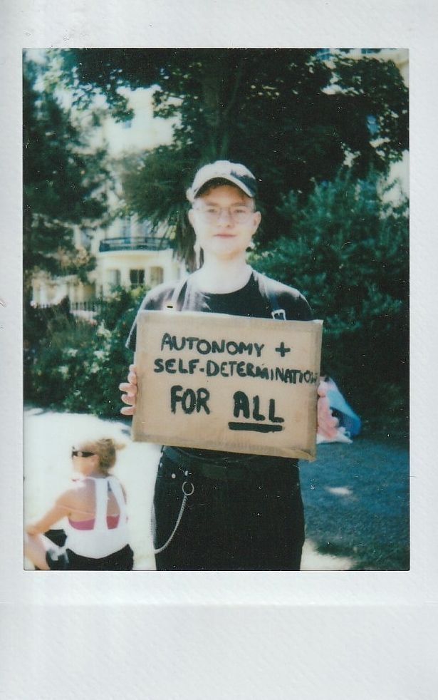 A person holds a cardboard sign stating "Autonomy + Self-Determination For All".