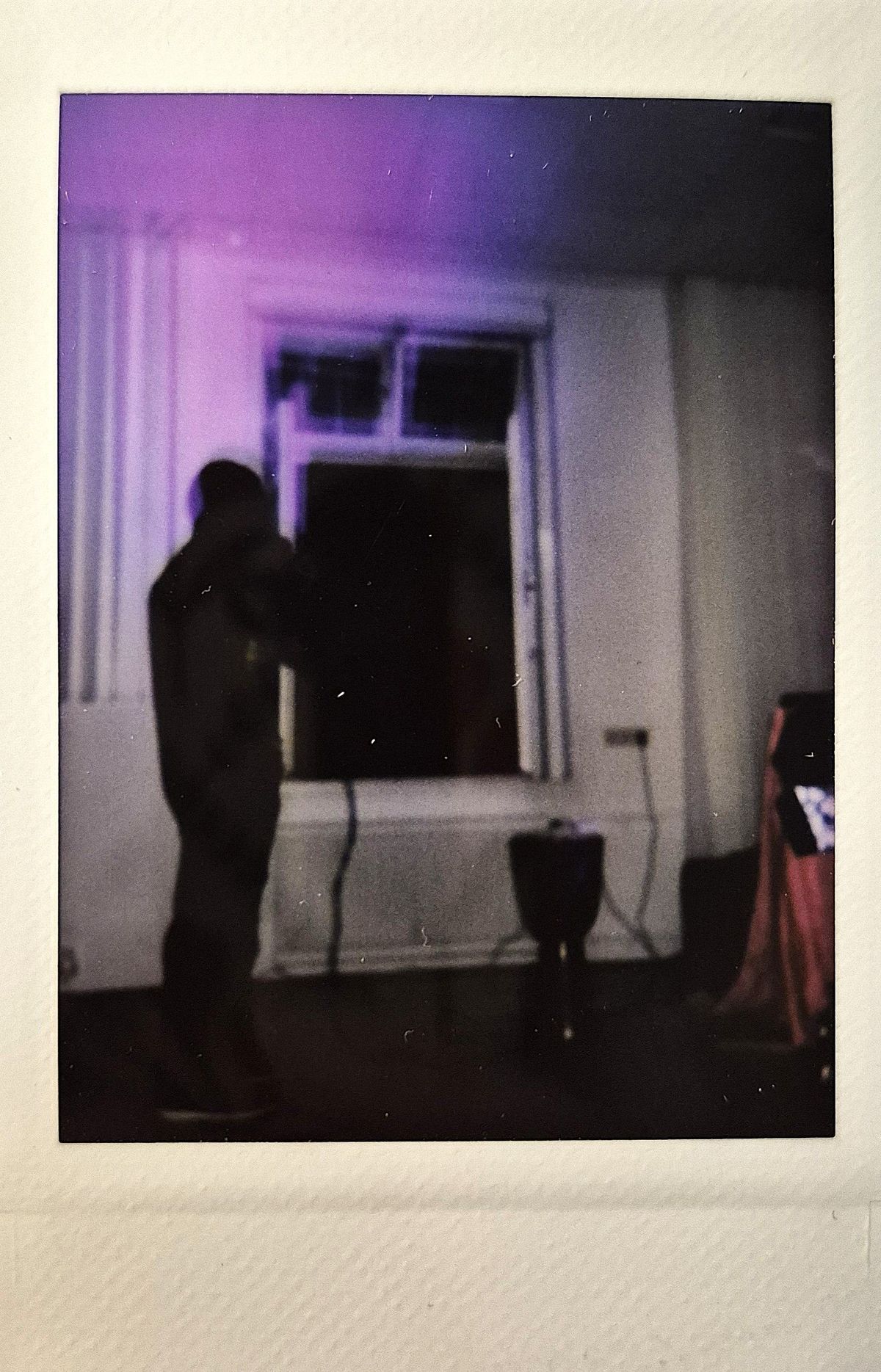 A silhouette stands by a window in a dimly lit room with purple light casting a subtle glow.