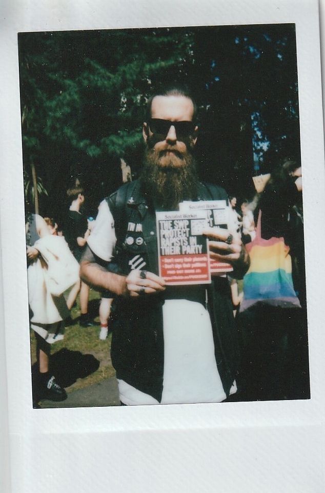 A bearded person in sunglasses holds a publication at an outdoor event, surrounded by people and trees.