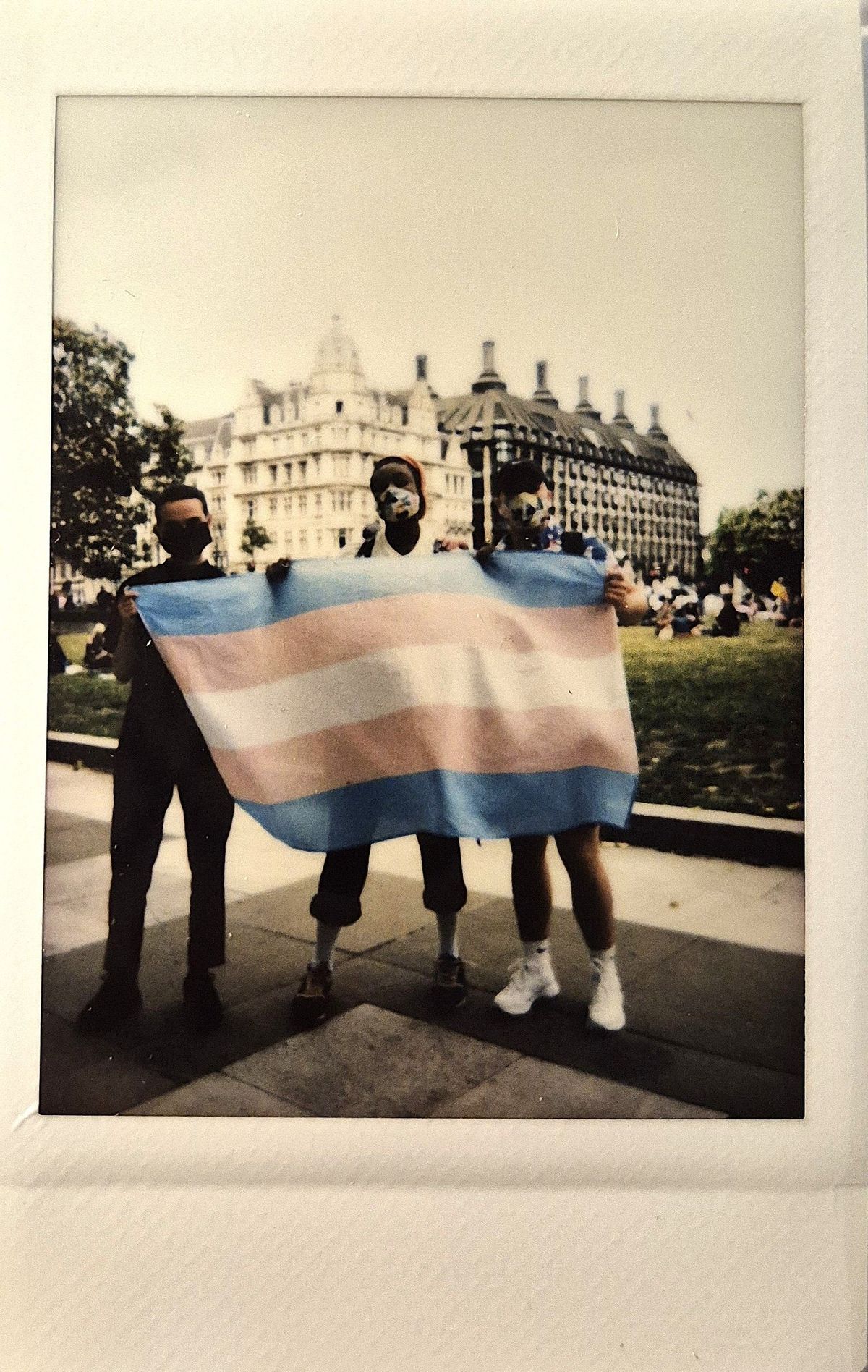 Three people hold a transgender pride flag outside near a large historic building.