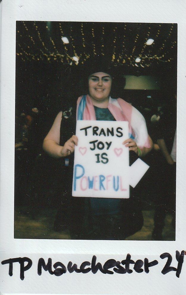 A person holds a sign saying "Trans Joy is Powerful," smiling under string lights.