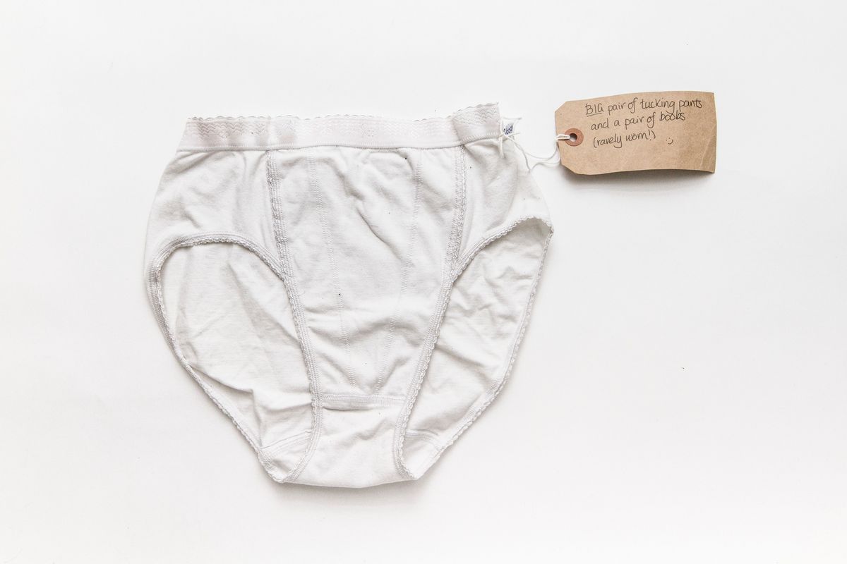 Pair of white tucking pants lying on a wooden surface. The cotton string of the tag is wrapped around the gusset.