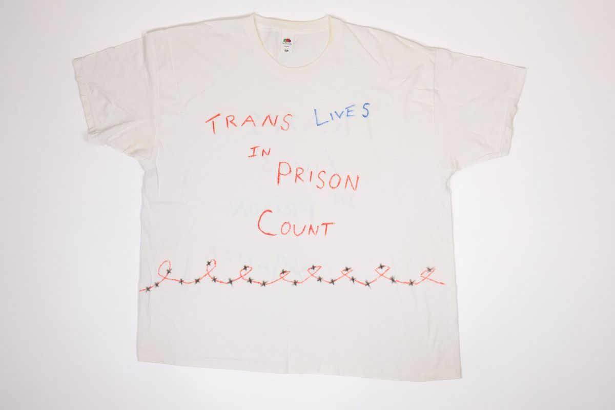 White T-shirt with handwritten text "TRANS LIVES IN PRISON COUNT" in red and blue and hand drawn red curly barbed wire with black 'x's.