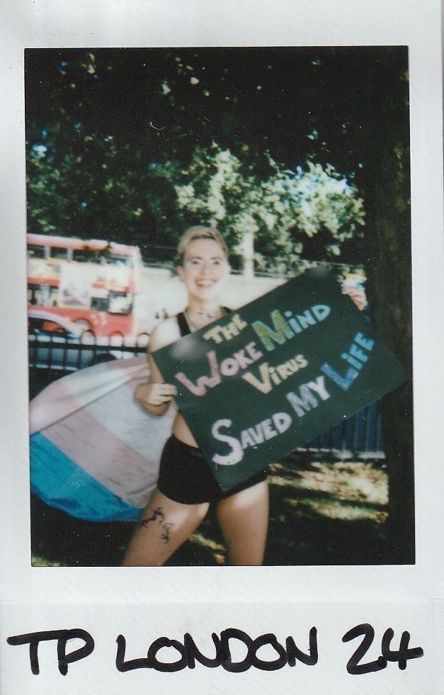 A person stands holding a colorful sign in a park, with a double-decker bus visible nearby.