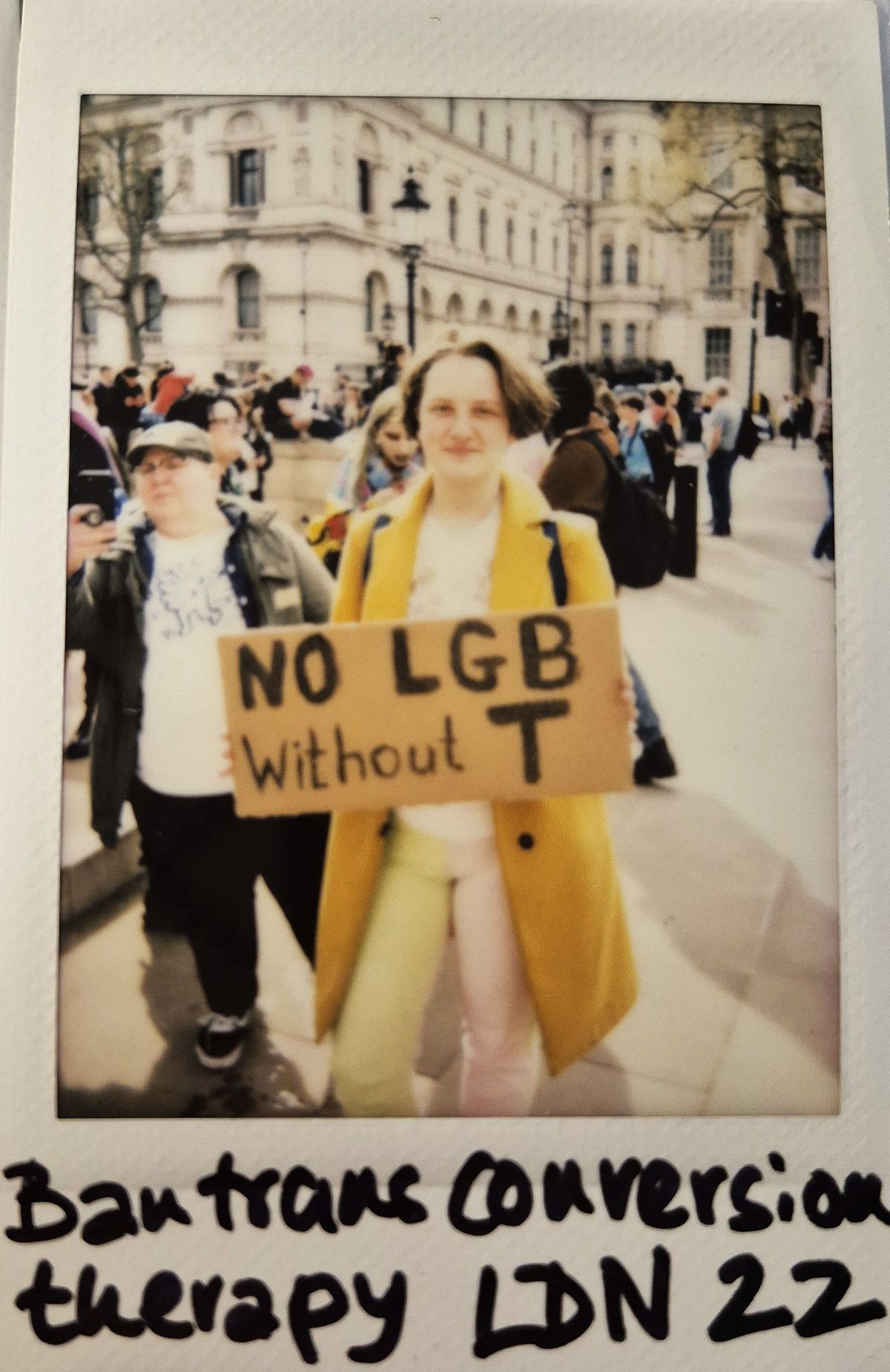 A person in a yellow coat holds a sign reading "No LGB without T" during a protest.