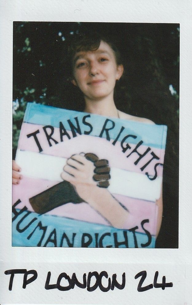 A person smiles, holding a sign that reads "Trans Rights Human Rights" with clasped hands.