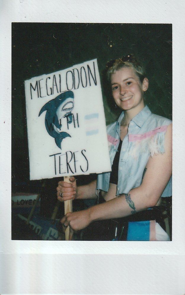 A person holds a sign reading "Megalodon With TERFS," featuring a drawing of a shark.