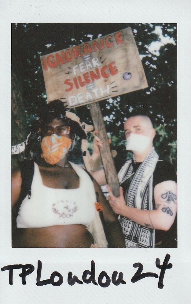 Two people, wearing masks, hold a sign that reads, "Ignorance + Fear + Silence = Death".