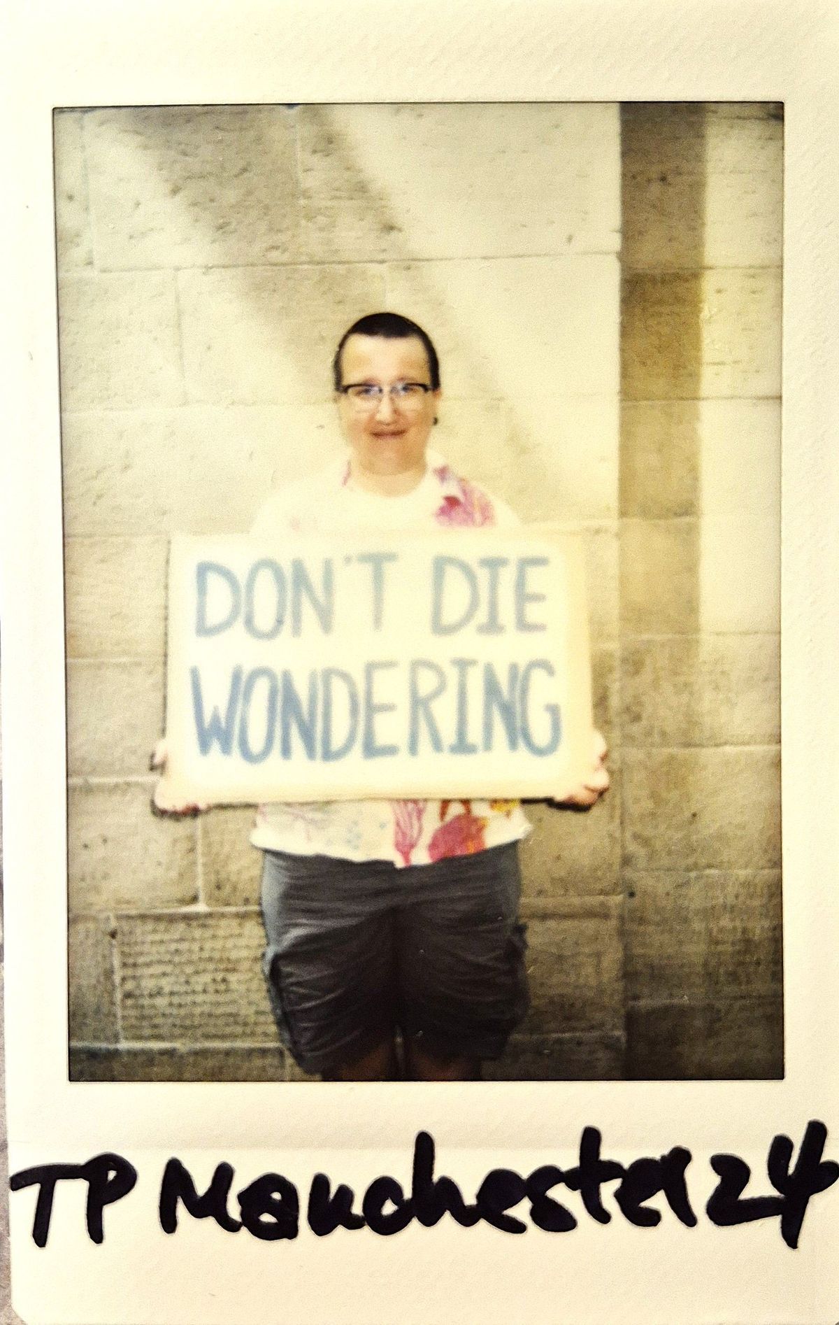 A polaroid of a person holds a sign reading "DON'T DIE WONDERING" against a stone wall backdrop.