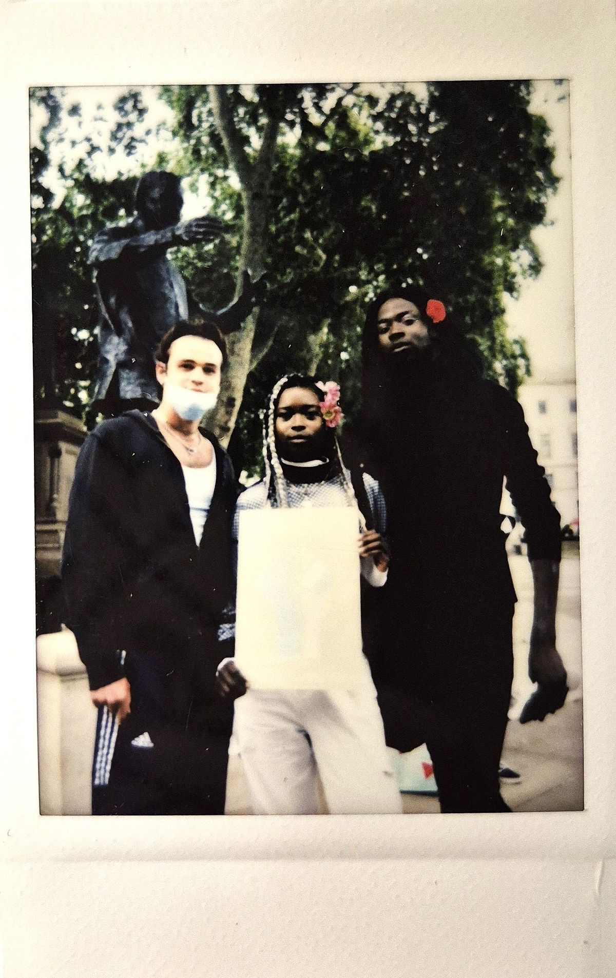 Three people stand together outdoors, two with flowers in their hair, in front of a statue.