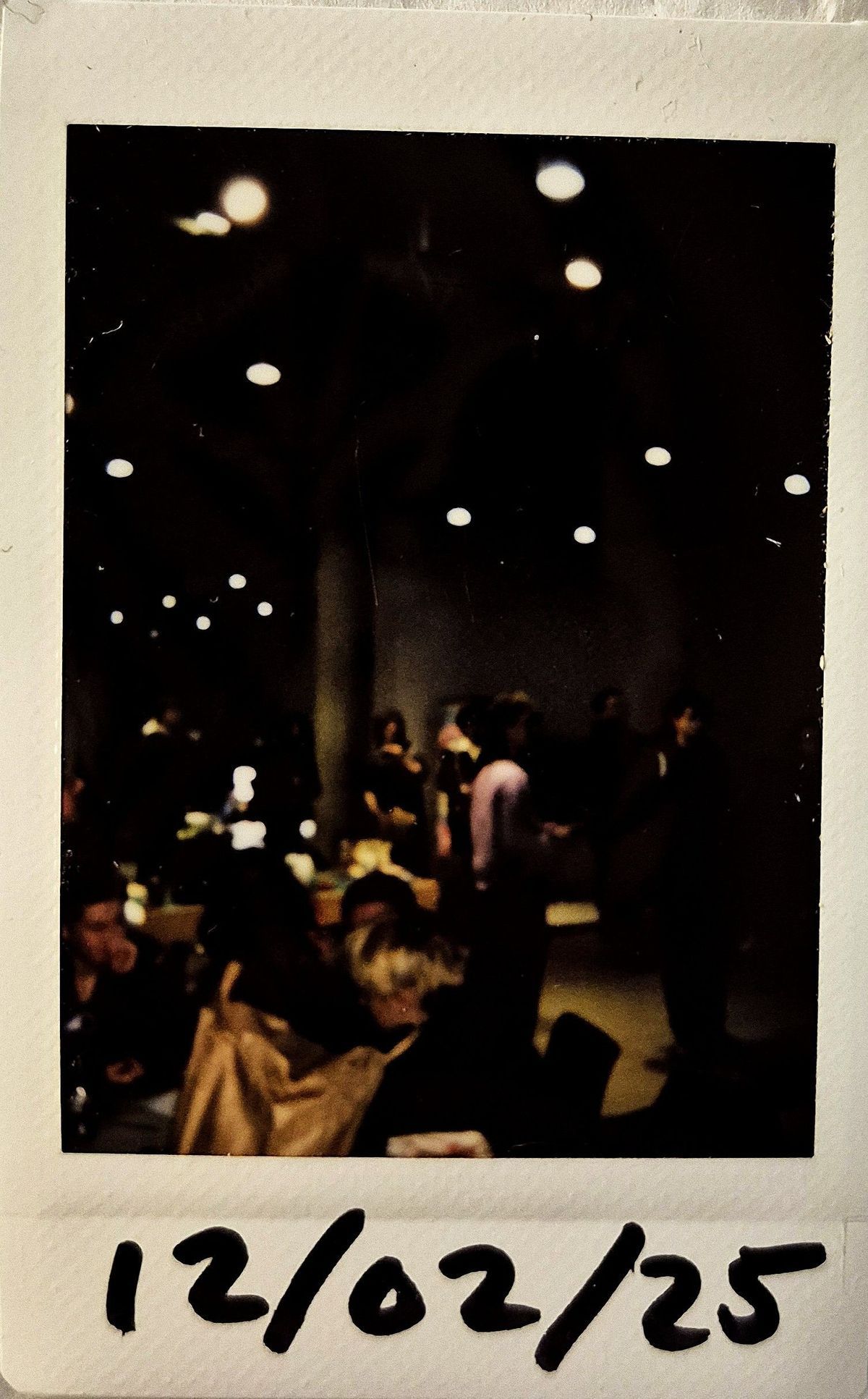 A dark lit polaroid which vaguely a room with lots of people and tables.