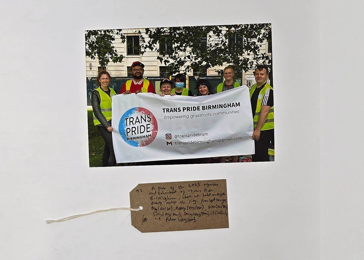 A group poses with a "Trans Pride Birmingham" banner, smiling and wearing yellow vests.