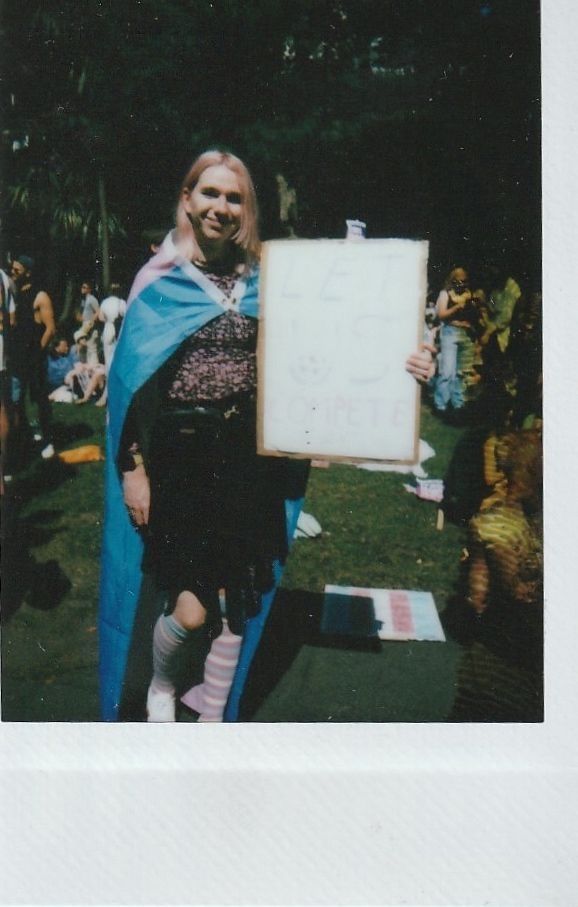A person draped in a colorful flag holds a sign stating "Let Us Compete".