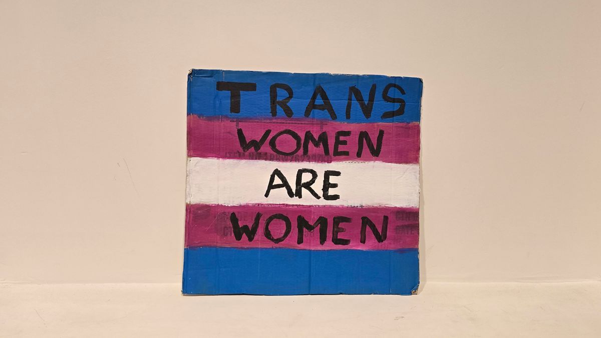 A placard with trans pride colors displays "Trans Women Are Women" in bold letters.