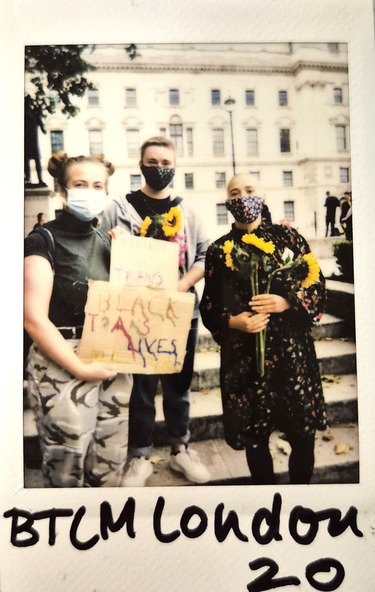 Three people wearing masks stand together holding sunflowers and a sign advocating for the protection of trans Black lives.