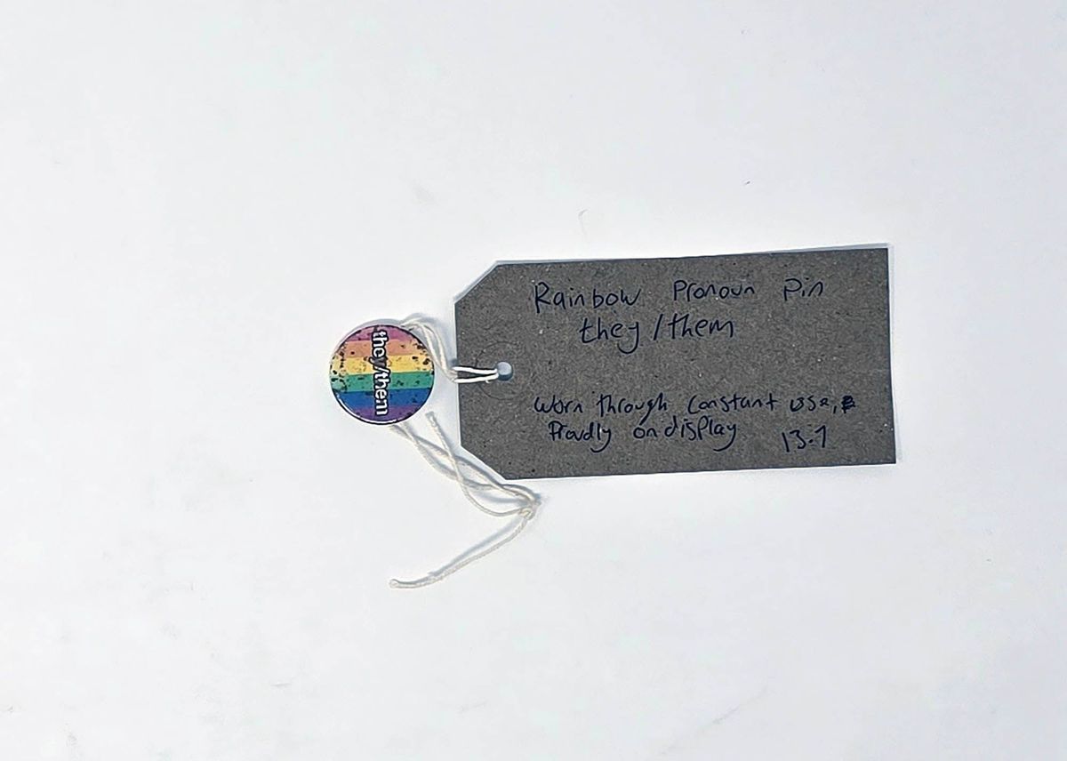 The image shows a rainbow pronoun pin labeled "they/them" with a tag describing its usage