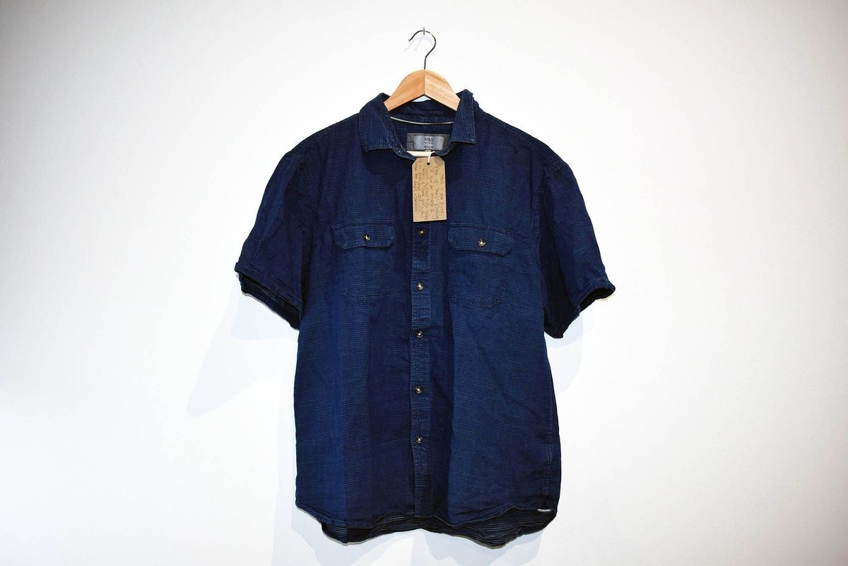 A blue short-sleeved shirt hangs on a wooden hanger against a plain white background.