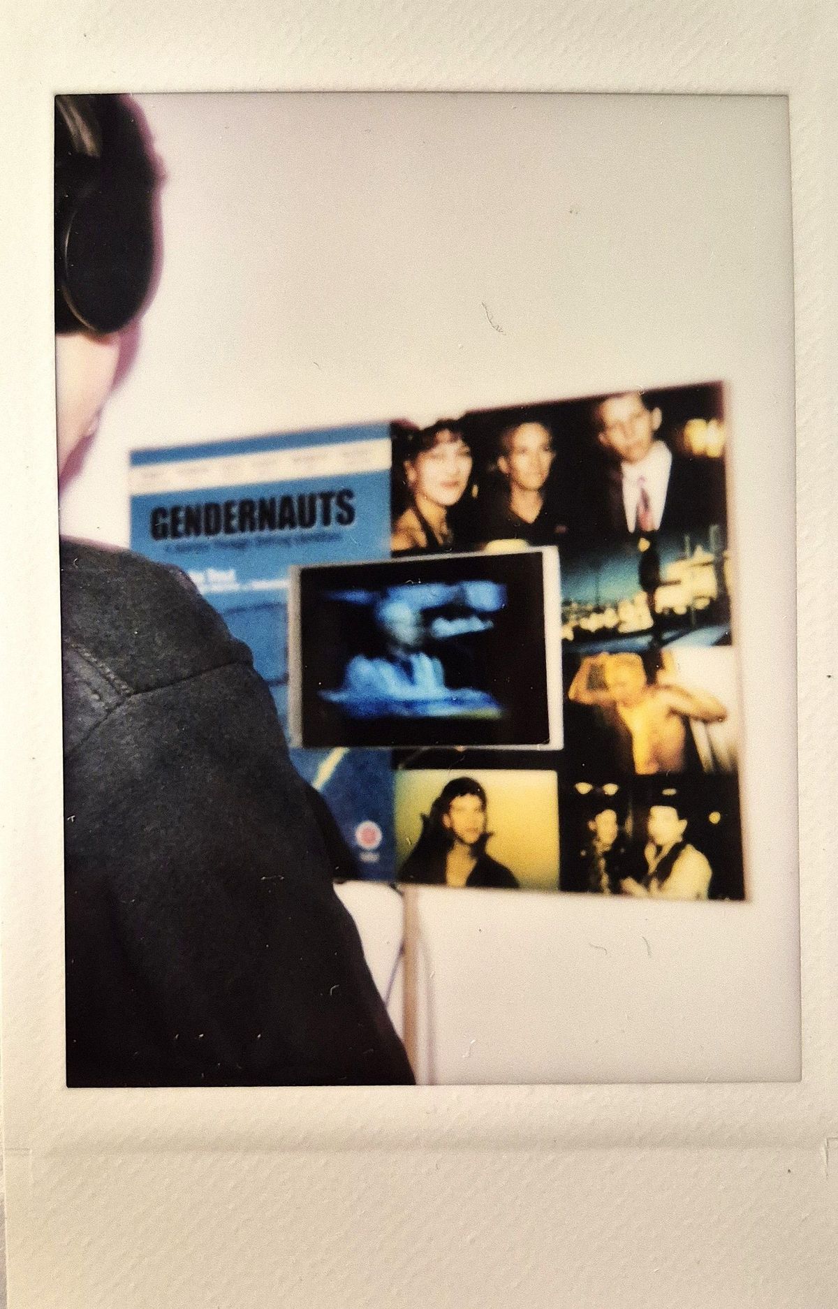 A person stands in front of a wall displaying posters and photographs, with a focus on "Gendernauts".