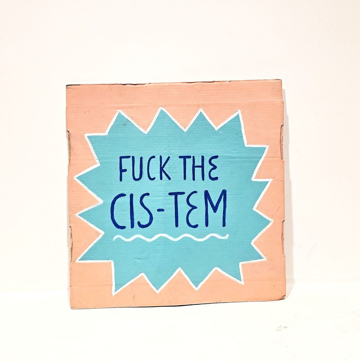 An orange cardboard placard with the slogan "FUCK THE CIS-TEM" in black lettering win a grey starburst.