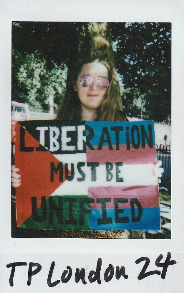 A person holds a colorful sign that reads "Liberation must be unified" beneath a tree.