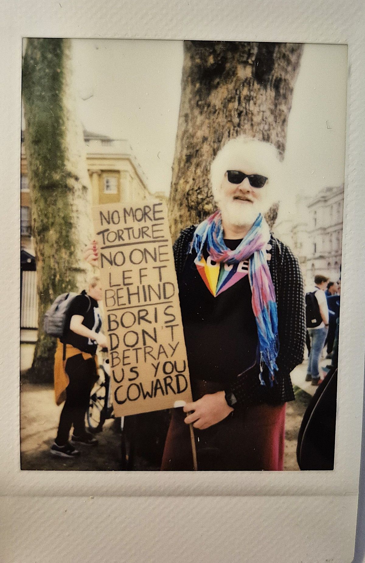 A person stands holding a protest sign which says "NO MORE TORTURE NO ONE LEFT BEHIND BORIS DON'T BETRAY US YOU COWARD".