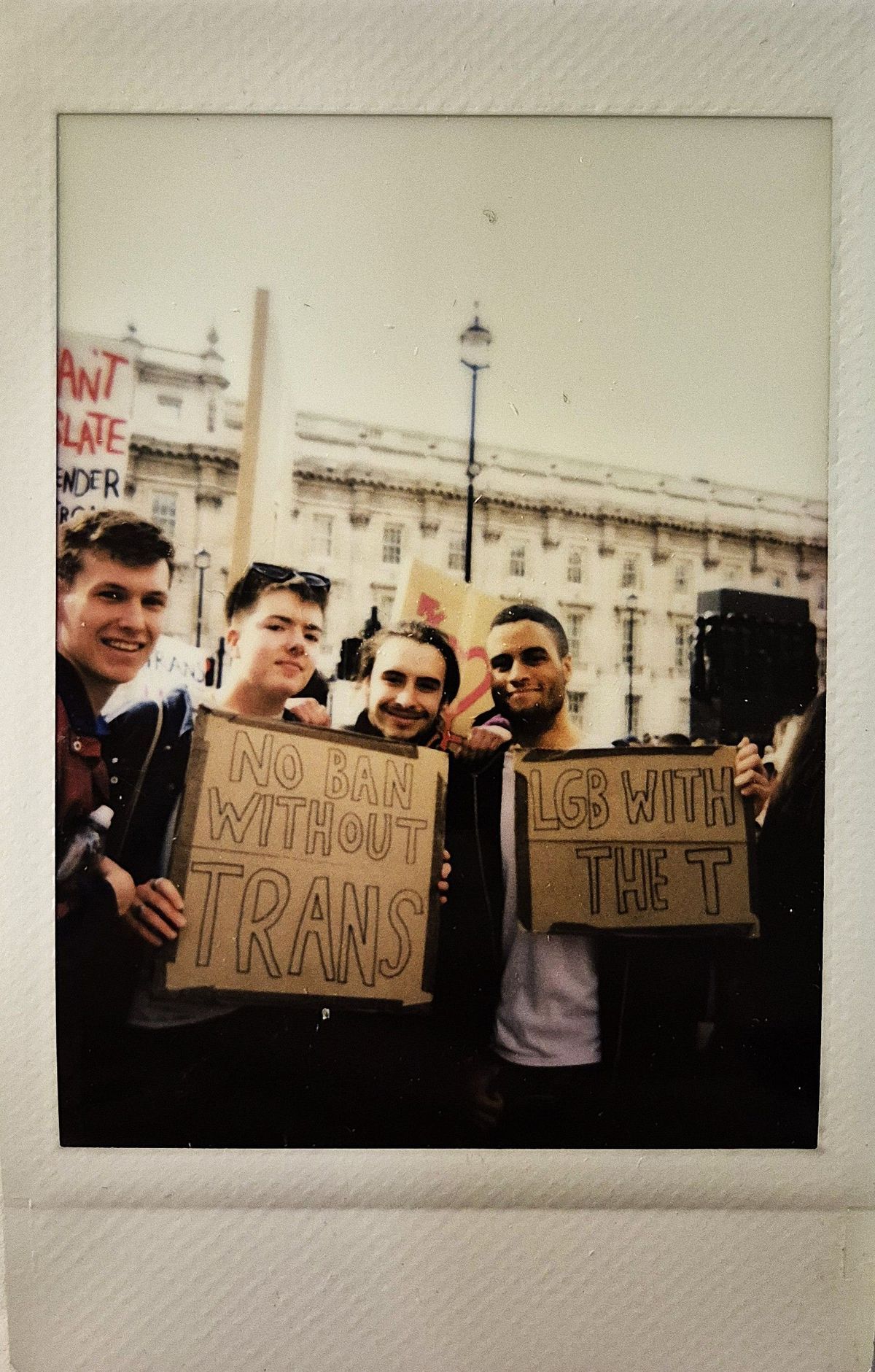 Four people are standing together, holding signs which says "NO BAN WITHOUT TRANS" and "LGB WITH THE T".