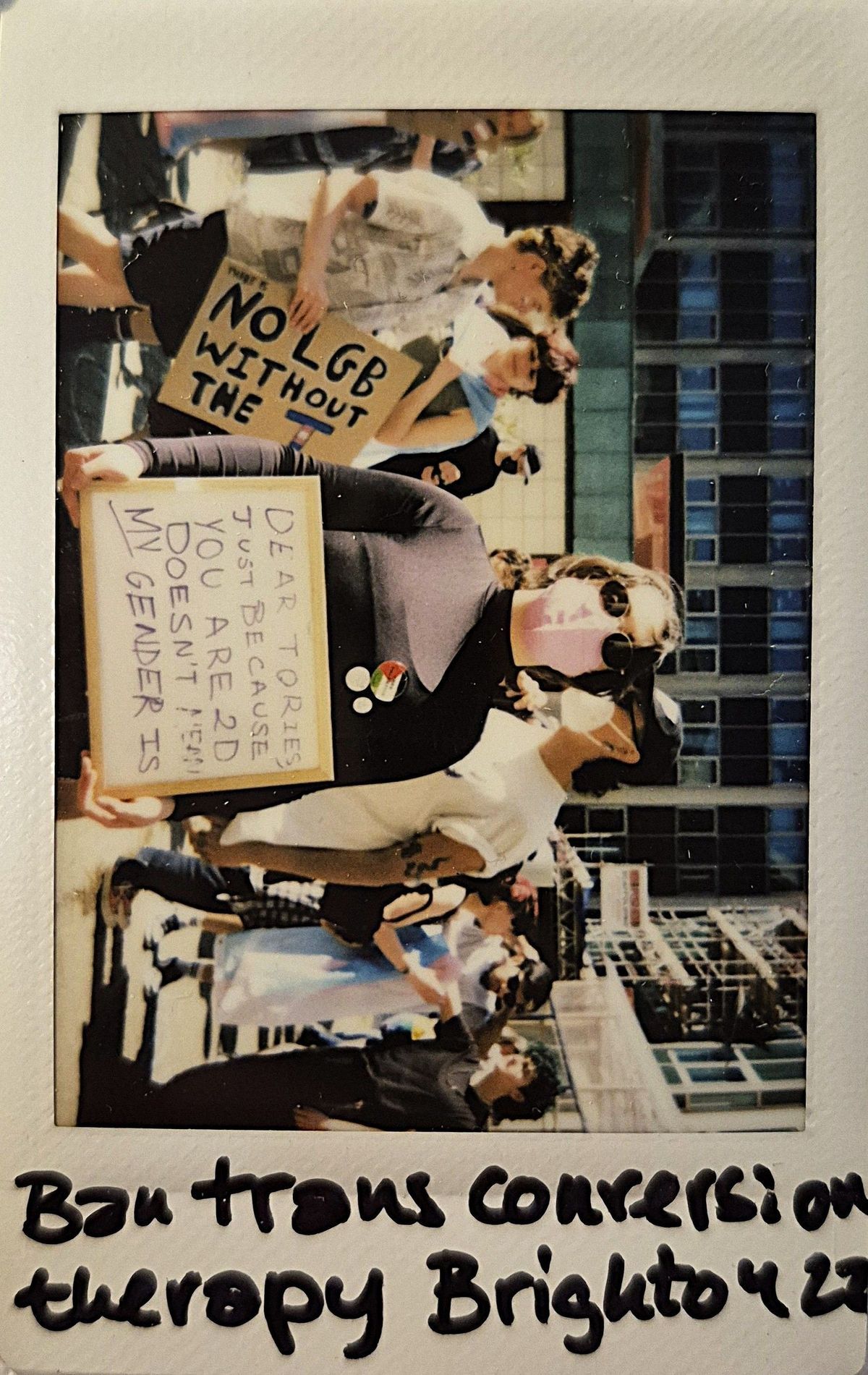 People are participating in an outdoor protest and one person is holding a sign which says "DEAR TORIES, JUST BECAUSE YOU ARE 2D DOESN'T MEAN MY GENDER IS".
