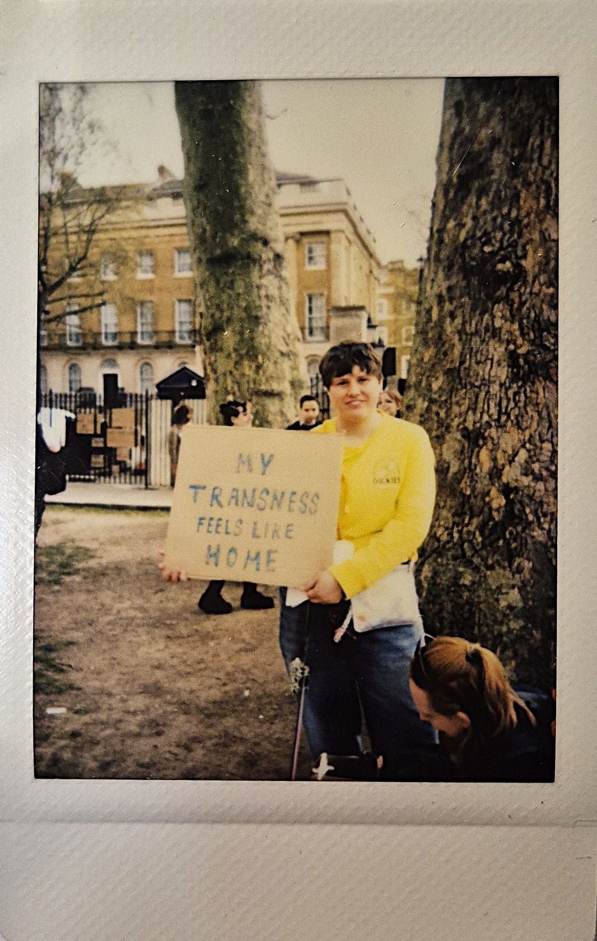 A person in a yellow shirt holds a sign reading "My transness feels like home".