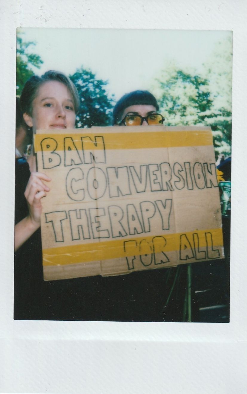 Two people hold a sign that reads "Ban Conversion Therapy For All" at an outdoor event.