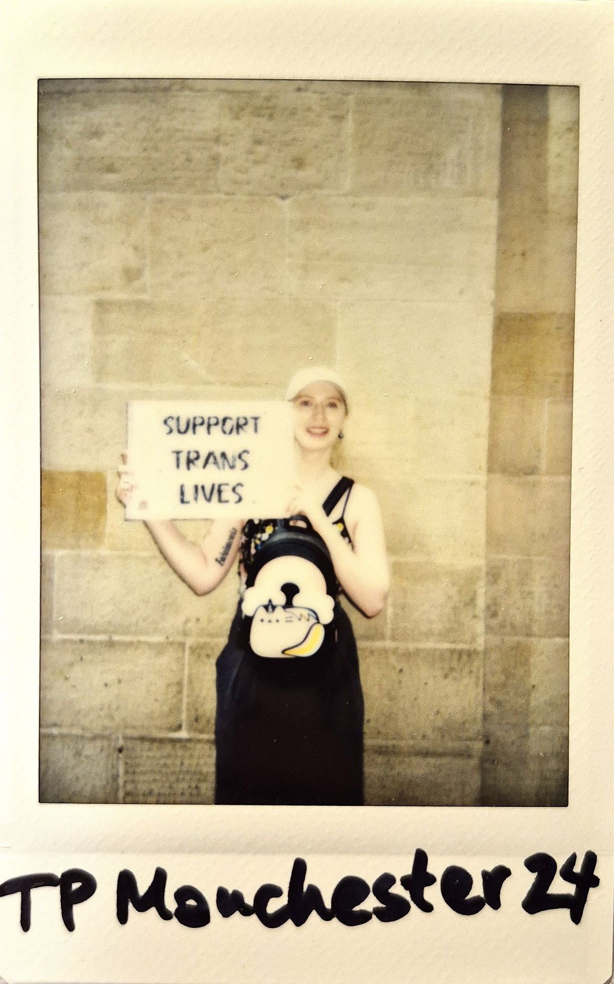 A polaroid of a person smiling holds a supportive sign reading "SUPPORT TRANS LIVES" against a stone wall background.