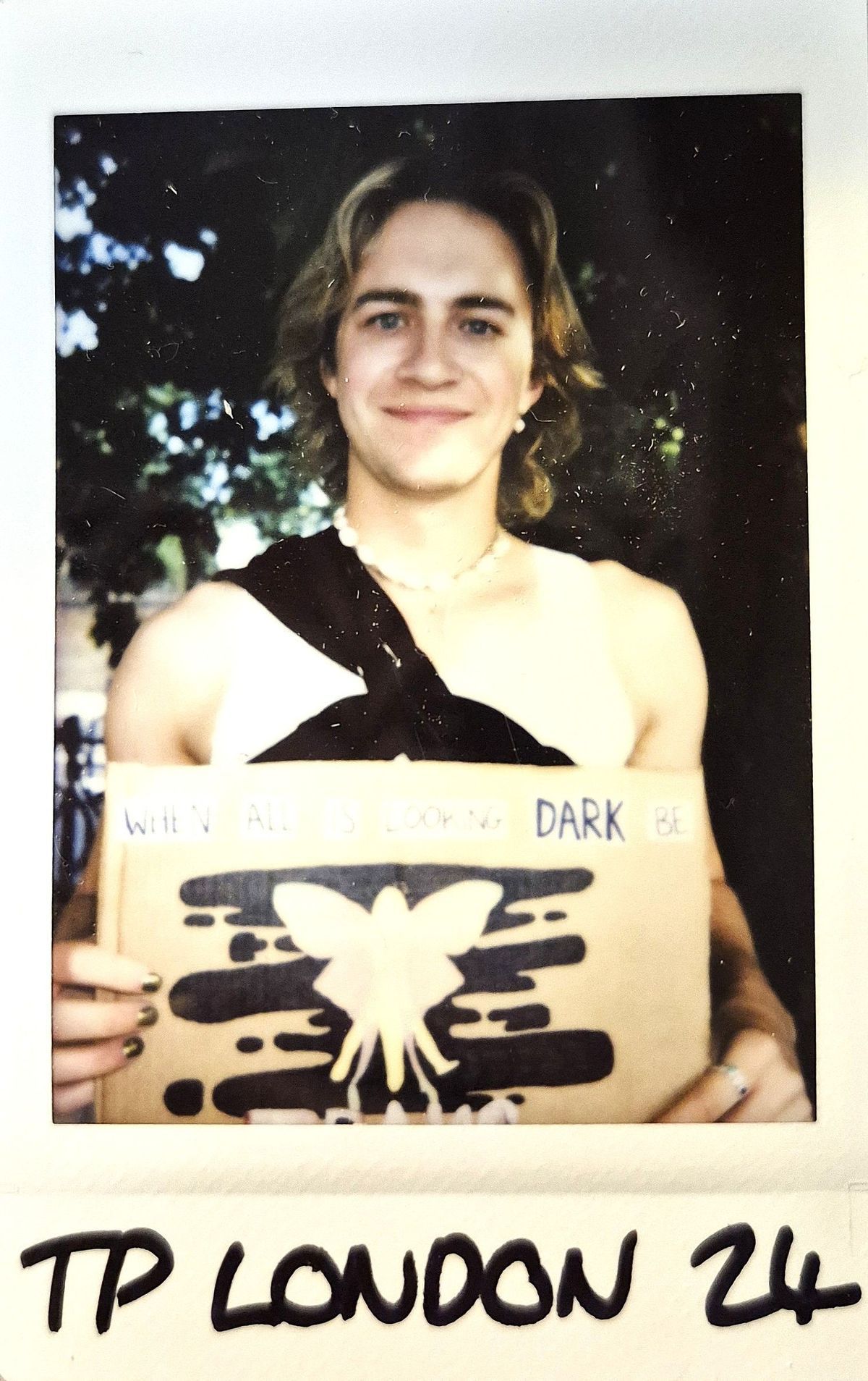 A smiling person holds a sign with an inspirational message, featuring a butterfly design, surrounded by trees.