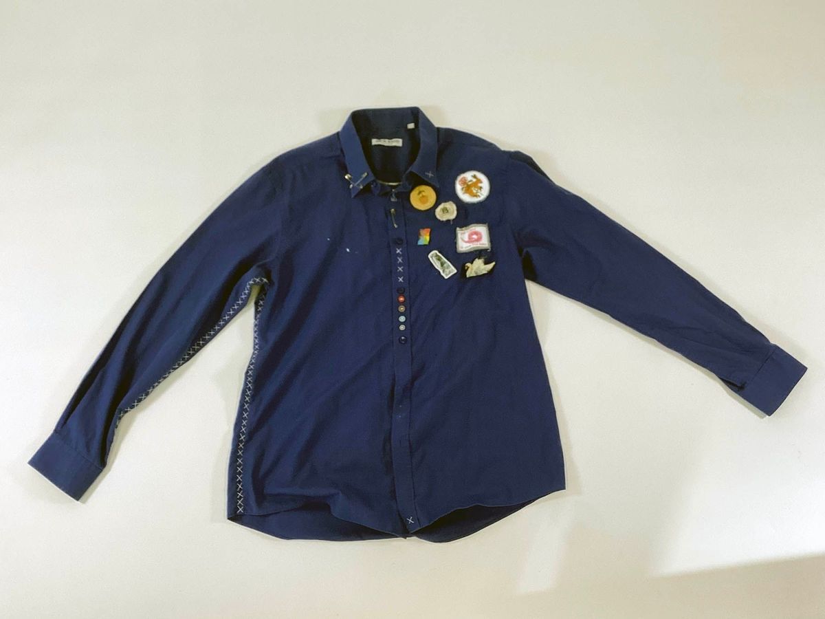 A dark blue button up shirt, with some colourful buttons. It has distinctive 'X' stitching up the side seams and down the arm seams. There are several patches sewn onto the left breast.