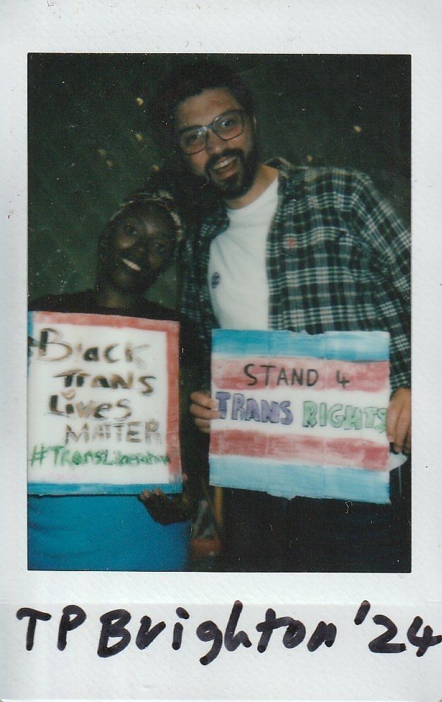 Two individuals hold signs supporting trans rights with smiles at an event marked TP Brighton '24.