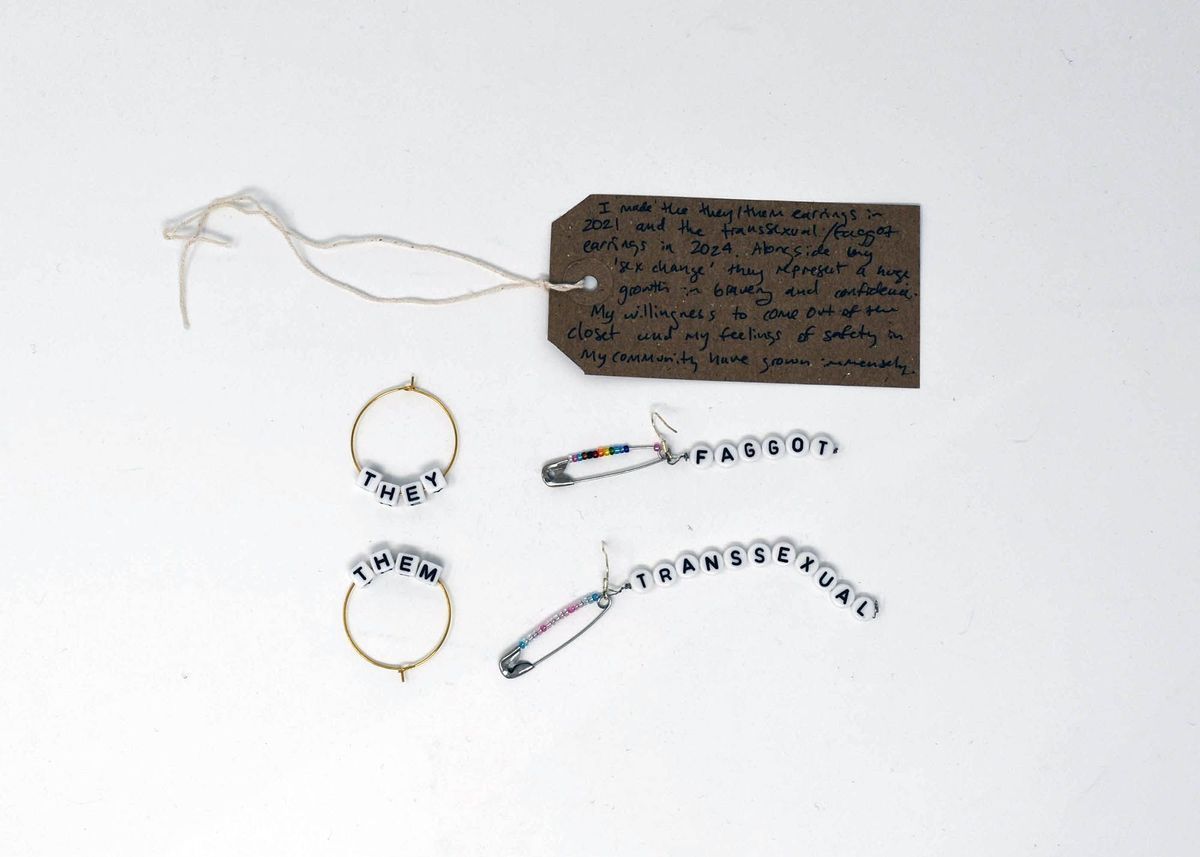 The image shows handmade earrings with pronouns and labels alongside a handwritten note attached with string.