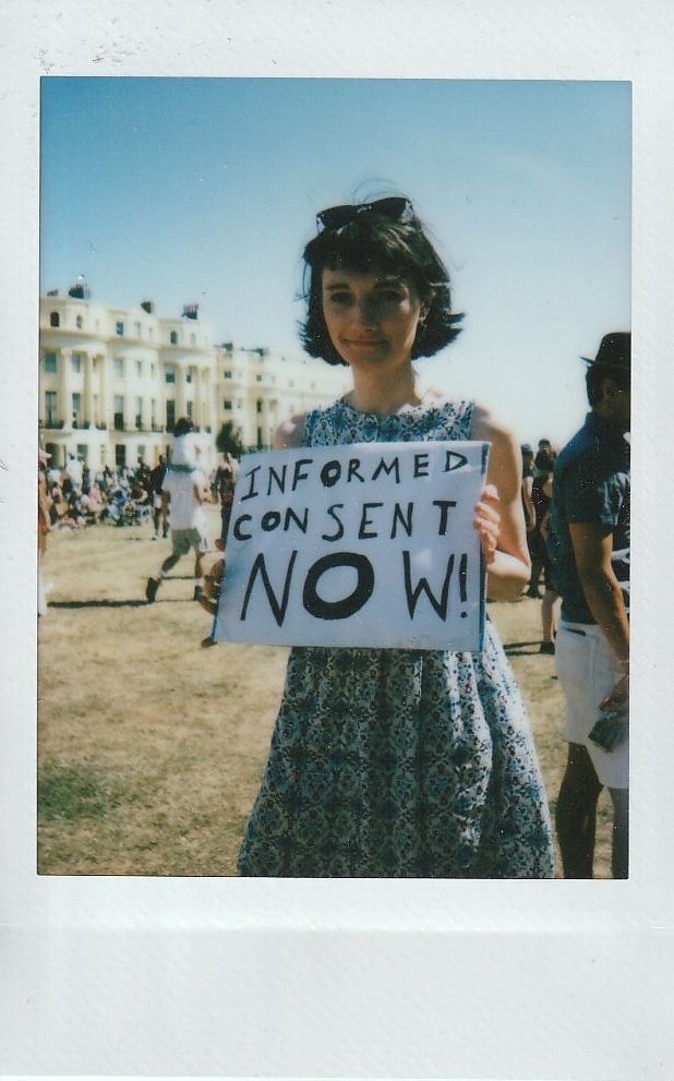 A person holds a sign that reads "Informed Consent Now!" at an outdoor gathering.