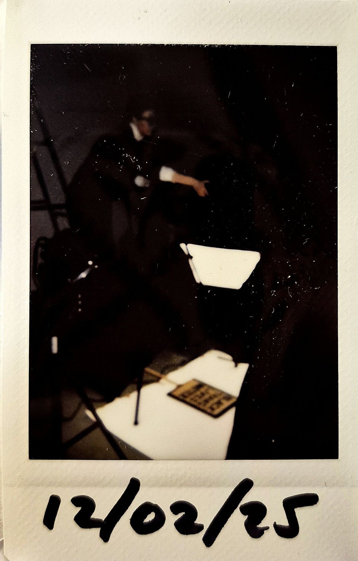 A dark lit polaroid which vaguely shows a photography lighting set up with a placard on the floor and someone stood over it.