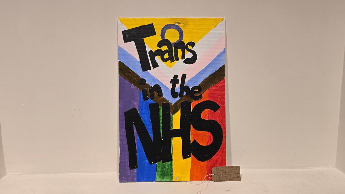 A placard which says “Trans in the NHS” with intertwined flag colors and bold lettering.