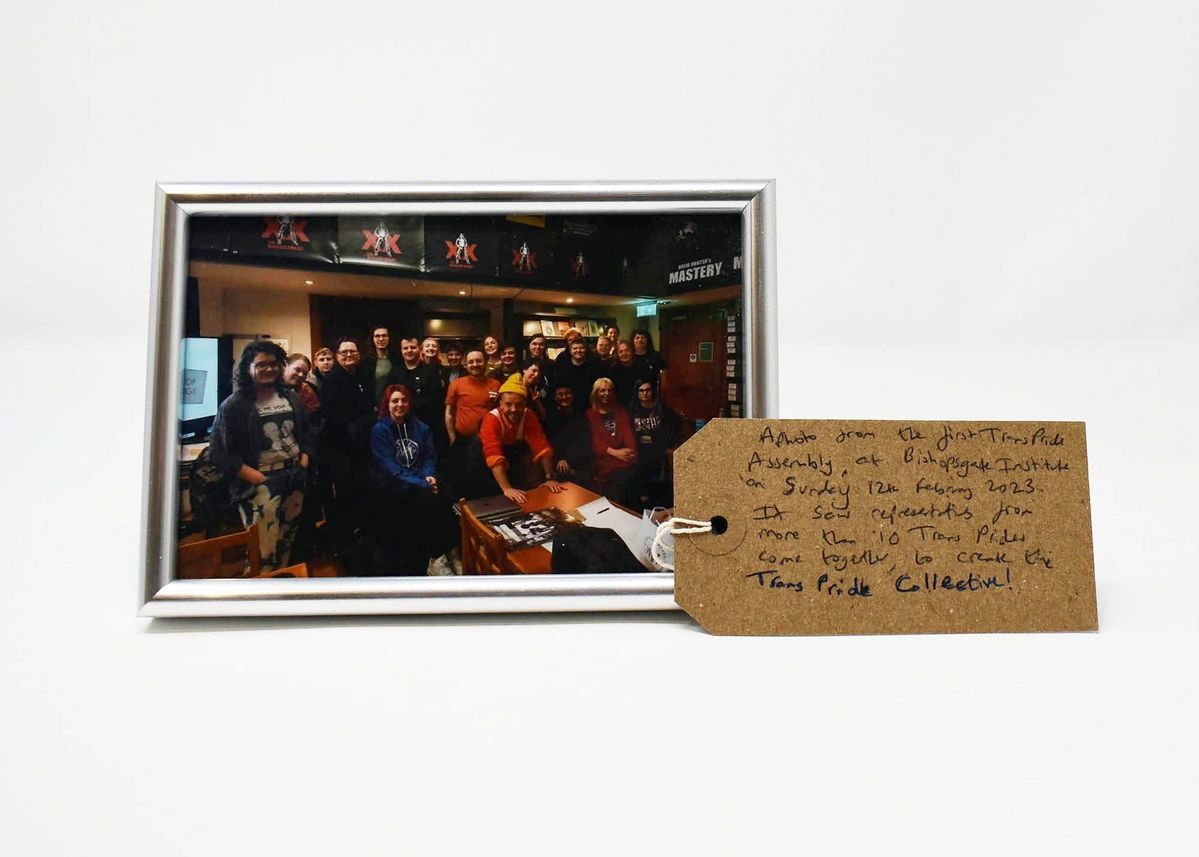 The image shows a group photo from a Trans Pride assembly with a handwritten tag detailing the event.