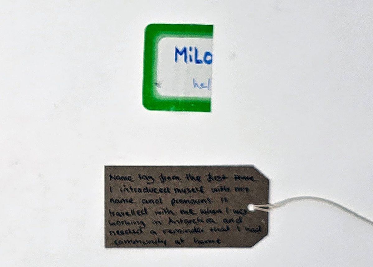 A folded over name label, white with a green border. The name "Milo" is visible, along with "he/". With a brown, handwritten luggage tag.