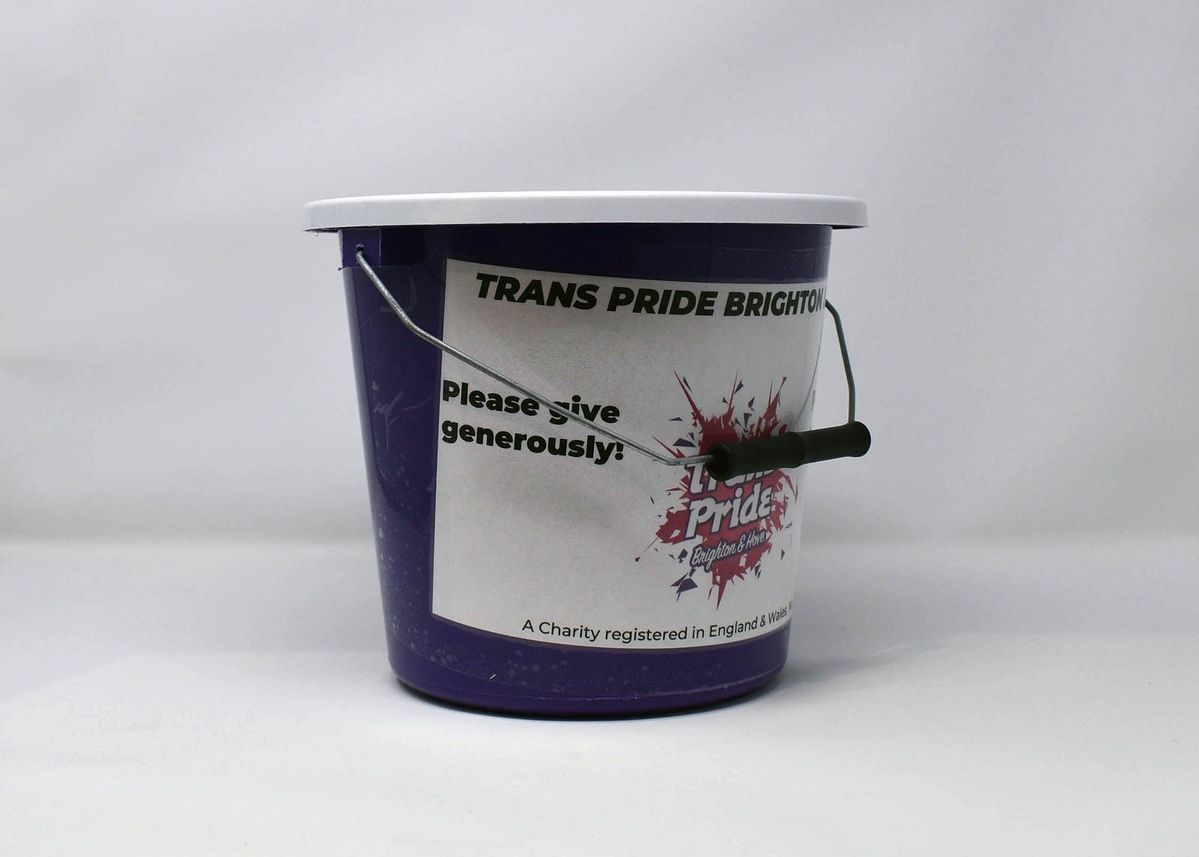 Purple coin collection bucket with white lid and white sticker wrapping around most of body. Several red and purple stickers are pasted on the lid and side with the "Trans Pride Brighton & Hove" logo.