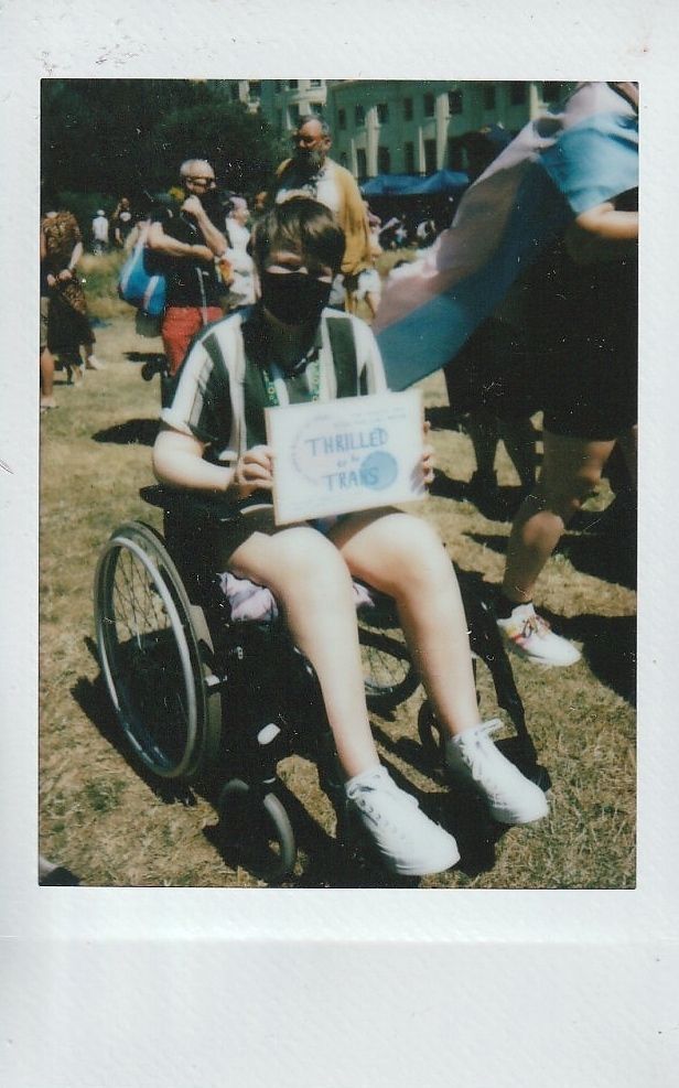 A person in a wheelchair holds a sign saying "THRILLED TRANS," surrounded by people outside.
