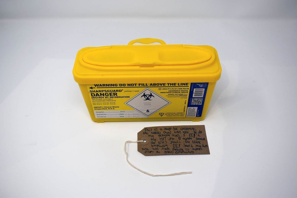 A bright yellow sharps box with a brown, handwritten luggage tag.