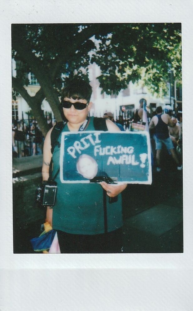 A person in sunglasses holds a sign which says "Priti Fucking Awful".