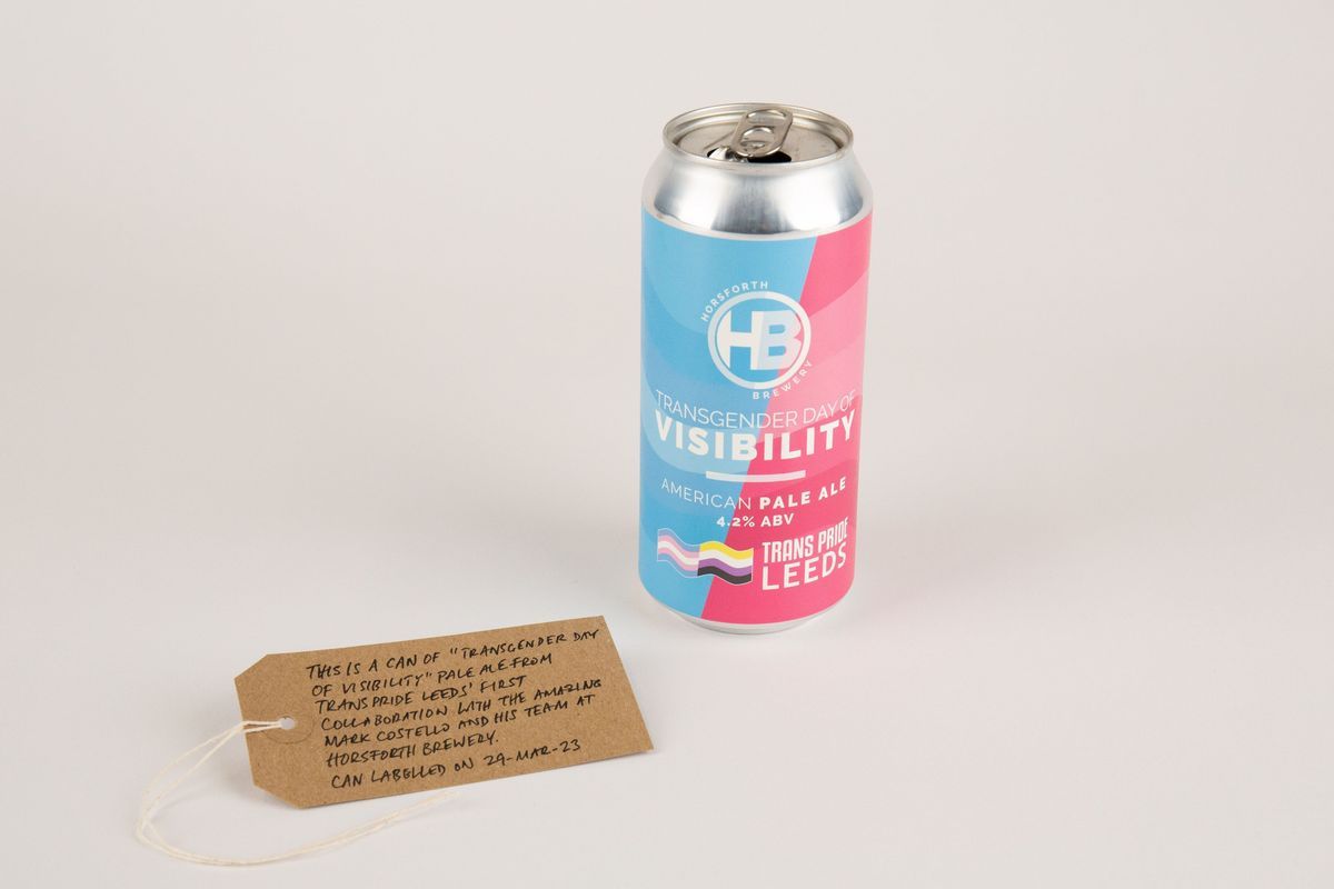 An empty can of "Transgender Day of Visibility" Pale Ale, brewed by Horsforth Brewery. The can is pink and blue and features the trans and non-binary flags. Also, a brown, handwritten luggage tag.