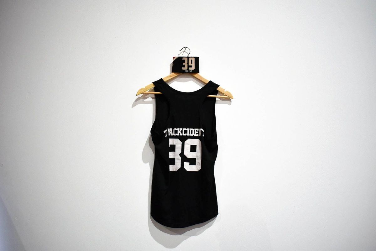 A black tank top with "TRACKCIDENT 39" hangs on a wooden hanger.
