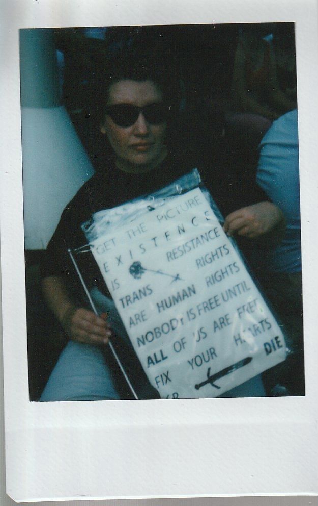 A person wearing sunglasses holds a placard sign which says "Get The Picture Existence Is Resistance Trans Rights Are Human Rights Nobody Is Free Until All Of Us Are Free Fix Your Hearts Or Die".