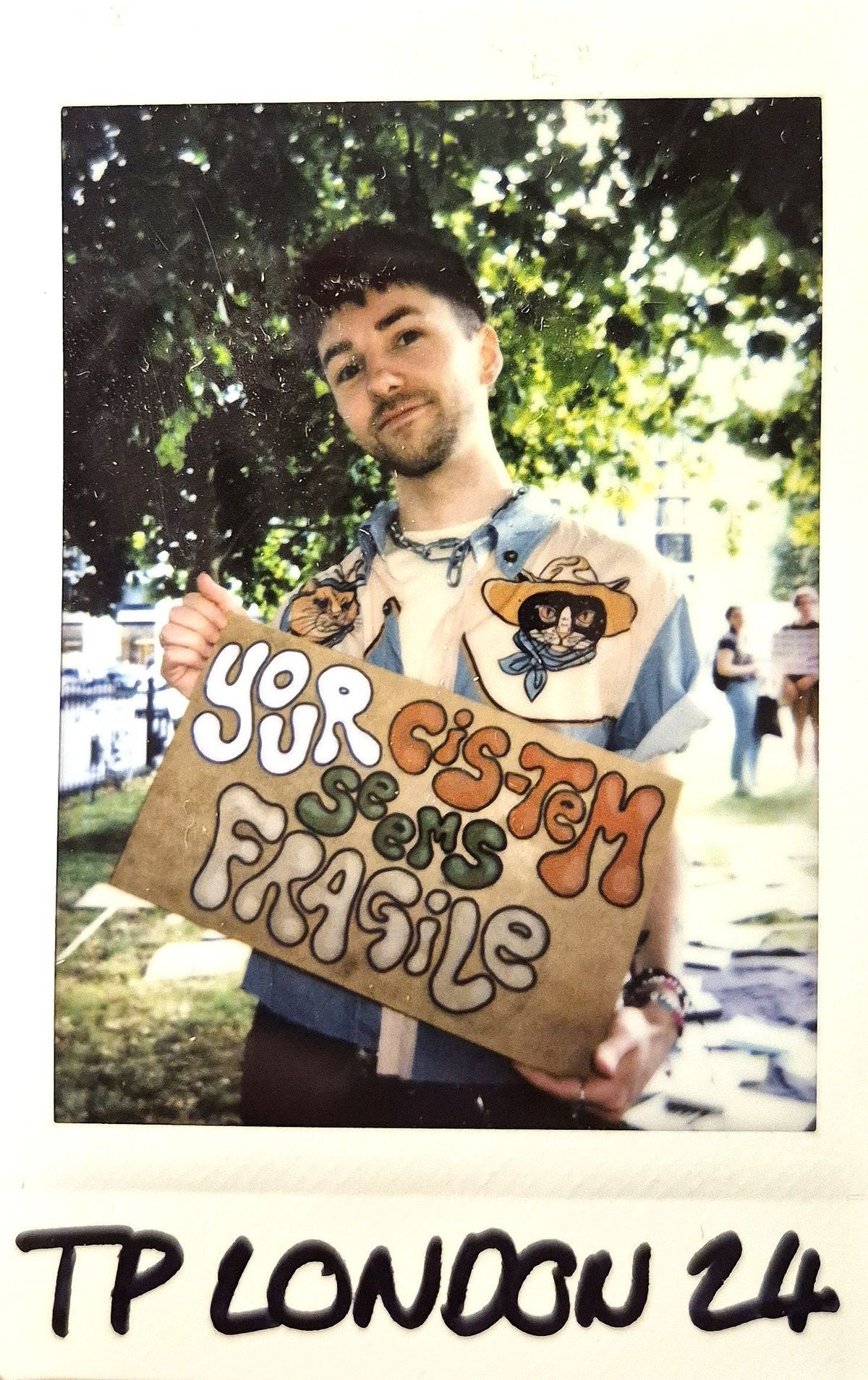 A person holds a sign saying, "YOUR CIS-TEM SEEMS FRAGILE," standing outside.
